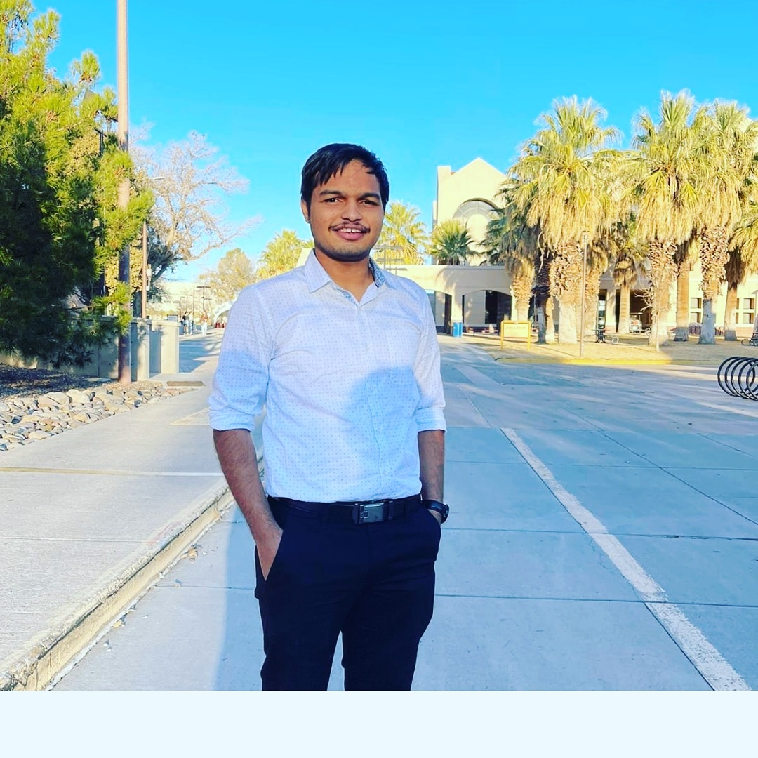 Adi Ghalsasi is pictured wearing a white button down shirt and black pants standing on the NMSU main campus in front of Zuhl Library. 
